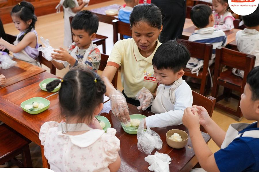 A teacher at Orange Crest Preschool helping children shape Bay Ben rice balls at a long wooden table, their hands in gloves and bowls of rice in front of them