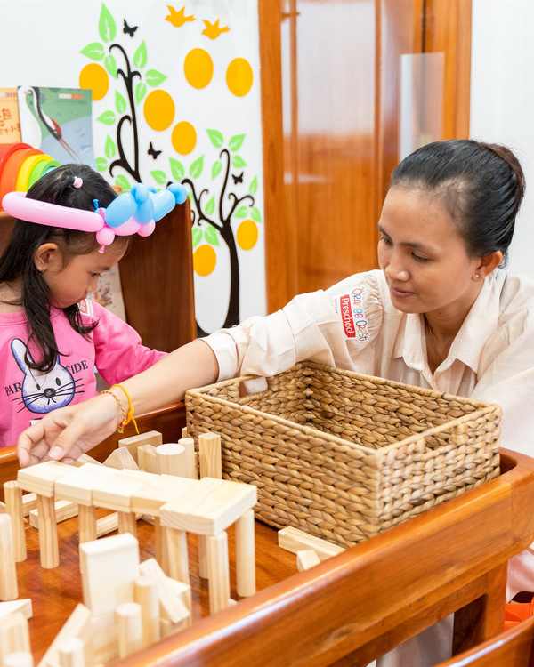 A teacher kneeling beside a child who is building with natural wooden blocks